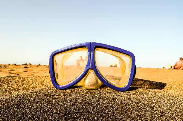 A pair of blue goggles is sitting on a sandy beach. The goggles are open and the lenses are clear. The scene is peaceful and relaxing, with the ocean in the background